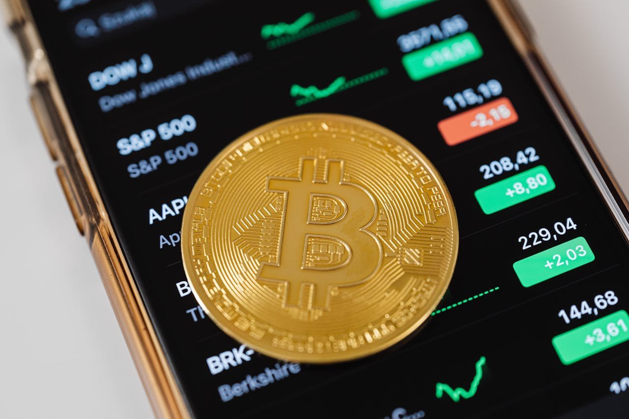 A close-up of a Bitcoin coin placed on a mobile device displaying stock market trading data.